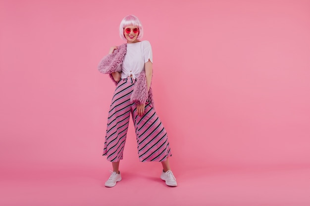 Full-length portrait of confident young woman in trendy pants. Indoor shot of inspired lady in peruke and sunglasses isolated on bright pink wall
