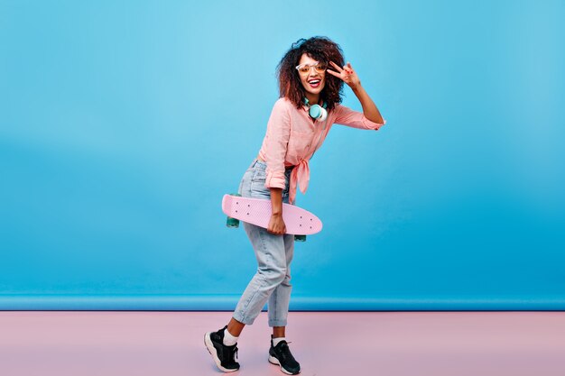 Full-length portrait of curly mulatto girl in black sneakers posing with peace sign after skating. Indoor photo of excited african lady with skateboard standing in front of blue wall with smile.