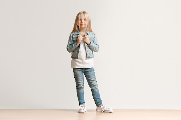 Foto gratuita ritratto integrale della ragazza carina ragazzino in jeans alla moda vestiti che guarda l'obbiettivo e sorridente, in piedi contro il muro bianco dello studio. concetto di moda per bambini