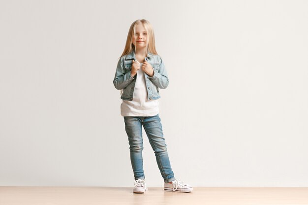 Full length portrait of cute little kid girl in stylish jeans clothes looking at camera and smiling, standing against white studio wall. Kids fashion concept