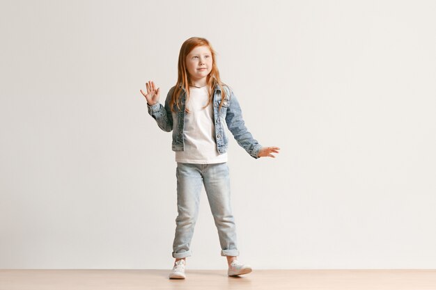 Full length portrait of cute little kid girl in stylish jeans clothes looking at camera and smiling, standing against white studio wall. Kids fashion concept