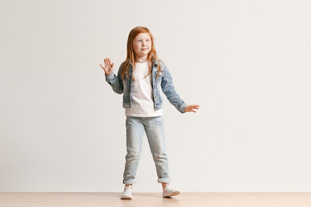 Полный портрет милой маленькой девочки в стильной джинсовой одежде, смотрящей на камеру и улыбающейся, стоящей на белой стене студии. Концепция детской моды