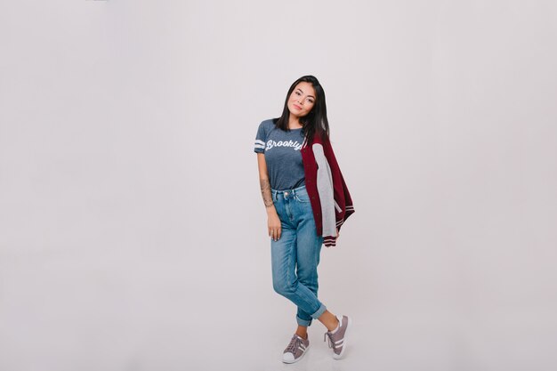 Full-length portrait of dark-haired female model in denim pants. Pretty brunette girl in jeans and trendy t-shirt posing