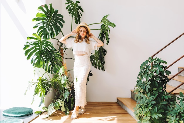 Full-length portrait of dreamy girl in white sneakers and skirt standing with hands up in shadow of big green flower