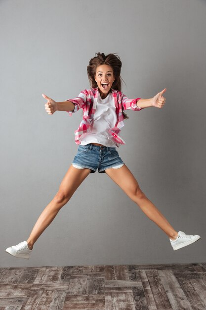 Full-length portrait of emotional jumping woman in casual wear, showing thumb up gesture,