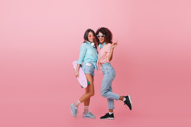 Full-length portrait of enchanting girl in cute socks and blue shirt standing near african female friend. black-haired lady with skateboard posing with mulatto young woman in jeans.