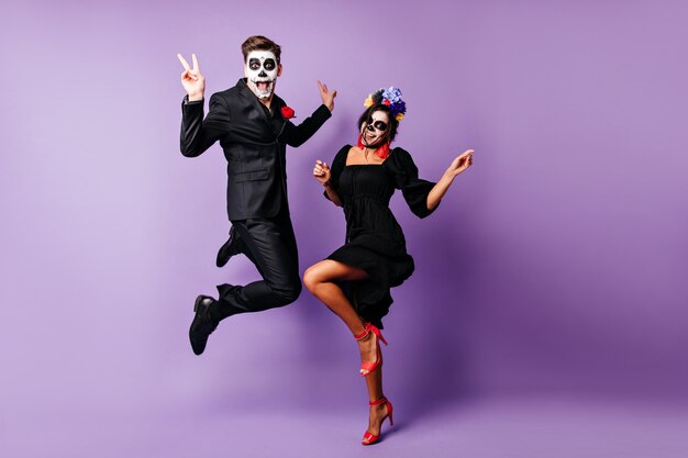 Full-length portrait of european couple dancing on purple background in zombie costumes. Funny young people fooling around at halloween event.