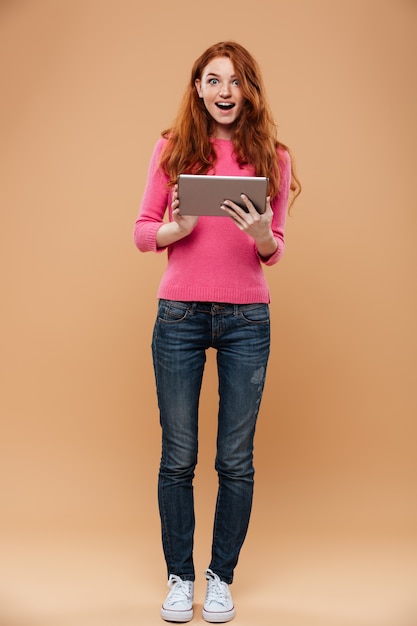 Full length portrait of an excited pretty redhead girl holding tablet