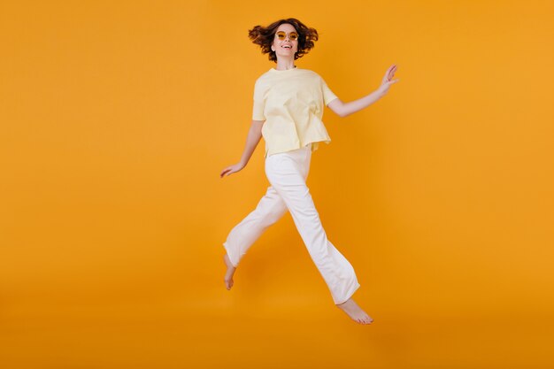 Портрет в полный рост радостной девушки в желтой футболке и белых штанах, бегущей по оранжевой стене. чудесная кавказская женщина танцует с удовольствием.