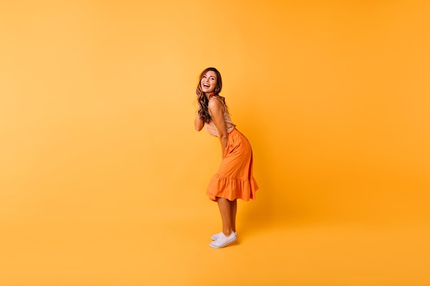オレンジ色のスカートでゴージャスな巻き毛の女の子のフルレングスの肖像画。黄色で踊るポジティブな女性のスタジオポートレート。