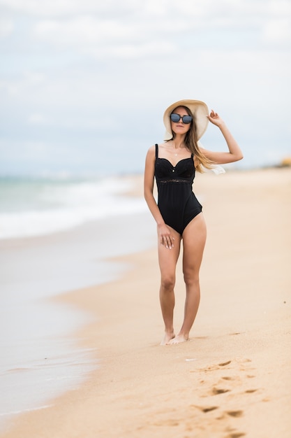 Full-length portrait of gorgeous young woman in straw hat walking on sandy beach