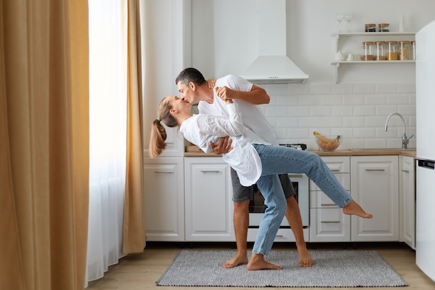 キッチンで一緒に踊るカジュアルな服を着て、夫が妻にキスをし、家で一緒に時間を過ごすのが幸せな幸せなカップルの全身像。