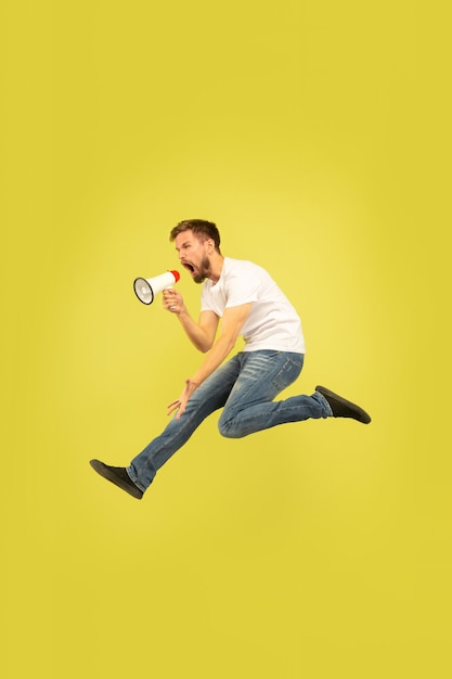 Full length portrait of happy jumping man isolated on yellow background. Caucasian male model in casual clothes. Freedom of choices, inspiration, human emotions concept. Calling with mouthpeace.