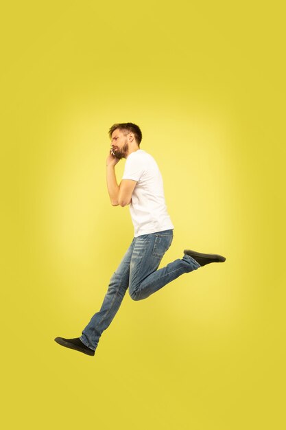 Full length portrait of happy jumping man isolated on yellow background. Caucasian male model in casual clothes. Freedom of choices, inspiration, human emotions concept. Hurry up, talking on phone.