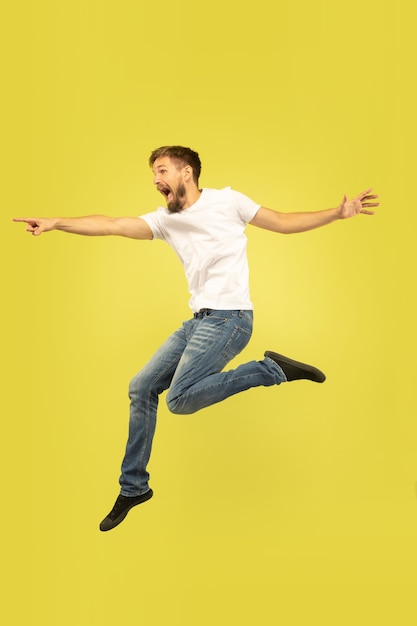 Full length portrait of happy jumping man isolated on yellow background. Caucasian male model in casual clothes. Freedom of choices, inspiration, human emotions concept. Pointing, choosing.