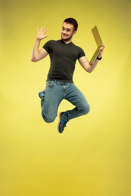 黄色の背景で隔離のガジェットを持つ幸せなジャンプ男の完全な長さの肖像画。現代のテクノロジー、選択の自由の概念、感情の概念。仕事や飛行中の楽しみのためにラップトップを使用する。
