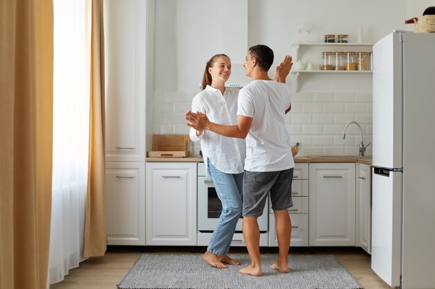 Полный портрет счастливой позитивной влюбленной пары, танцующей на кухне вместе, проводя время вместе дома, выражая романтические чувства.