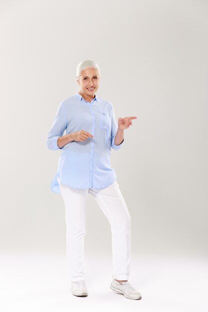 Full-length portrait of happy senior  gray-haired woman, standing and pointing with fingers