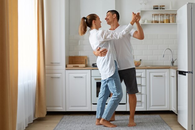 幸せな笑顔の夫と妻が明るい部屋で一緒に家で踊っているフルレングスの肖像画、キッチンセット、背景に冷蔵庫と窓、幸せなカップル。