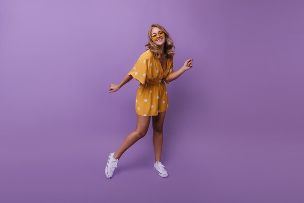 Full-length portrait of happy tanned girl in white sneakers. Portrait of pleased blonde woman dancing during portraitshoot on purple.