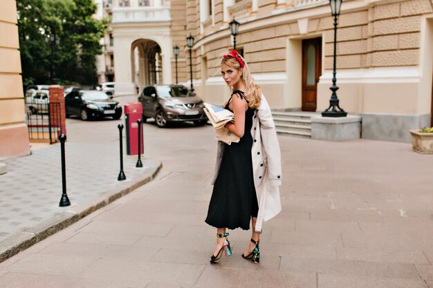 Full-length portrait of inspired blonde woman in black dress looks over shoulder and gently smiling in city square