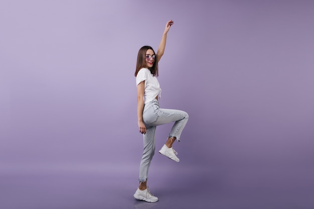 Full-length portrait of inspired female model in denim pants standing on one leg. brunette woman dancing with pleasure.