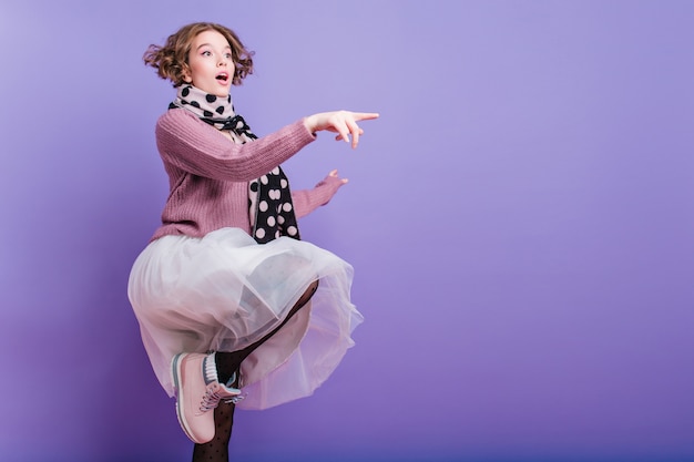 Full-length portrait of inspired girl in lush skirt posing on one leg on purple wall. graceful caucasian young woman in hat and long scarf having fun