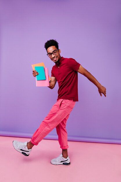 Full-length portrait of joyful international student dancing after exams. Smart african guy in pink pants standing with books.