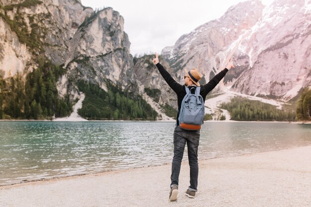 Full-length portrait of joyful man wearing hat having fun near river in cloudy morning and looking at Alps