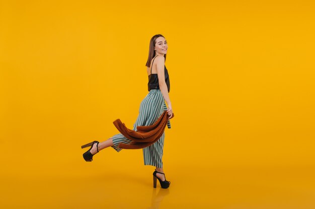 Полнометражный портрет смеющейся женской модели в туфлях на высоком каблуке с удовольствием. Фотография стройной стильной дамы в помещении.