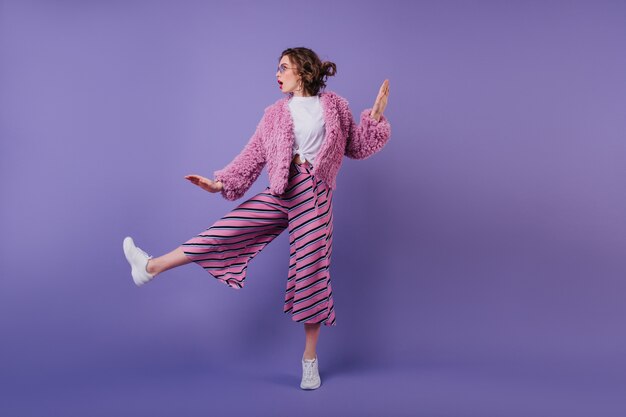 Портрет в полный рост привлекательной кудрявой женщины, танцующей в полосатых штанах. модная брюнетка девушка прыгает на фиолетовую стену.