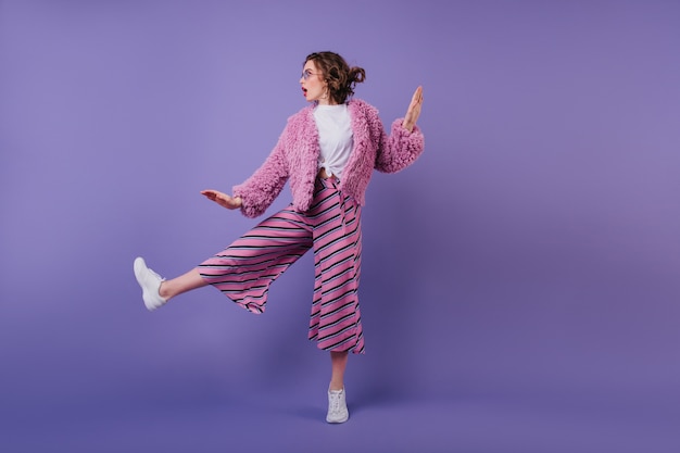 Full-length portrait of lovable curly woman dancing in striped pants. fashionable brunette girl jumping on purple wall.