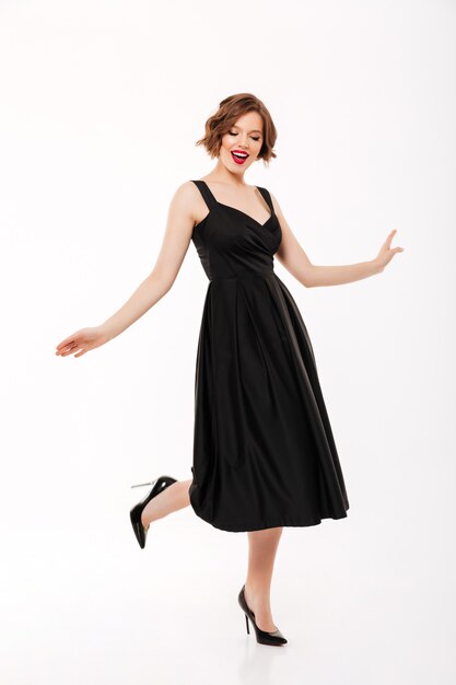 Full length portrait of a lovely girl dressed in dress pointing finger at a gift box while standing isolated over pink background