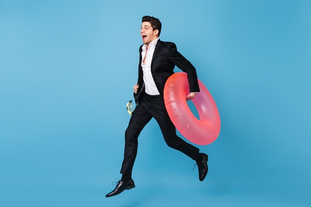 Free photo full-length portrait of man in business outfit running on blue space with pink inflatable circle.