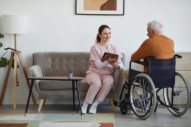 車椅子​で​年配​の​男性​と​話し​、​退職後​の​家​、​コピー​スペース​で​デジタル​タブレット​を​使用して​笑顔​の​女性​看護師​の​完全な​長さ​の​肖像画