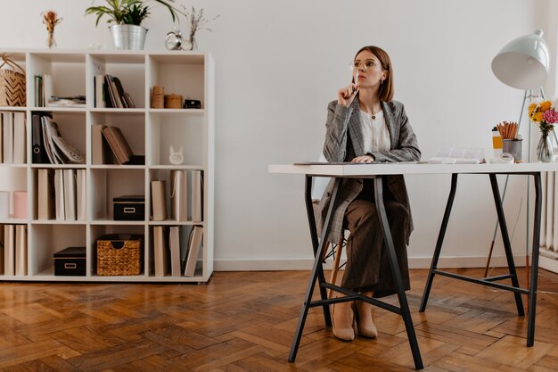 Full-length portrait of pensive business woman in trendy outfit sitting in minimalistic office.