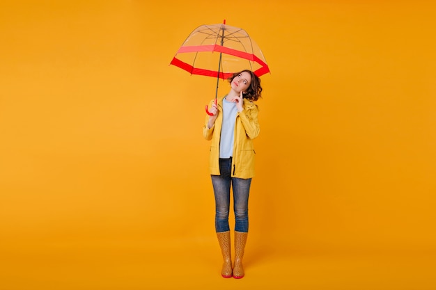 Full-length portrait of pensive romantic girl standing on yellow wall under red parasol. Studio shot of stylish female model in jeans and autumn shoes looking away while posing with umbrella.