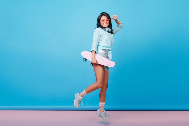 Full-length portrait of pleased latin lady in pink socks laughing and jumping. Active hispanic woman in sunglasses fooling around with skateboard.