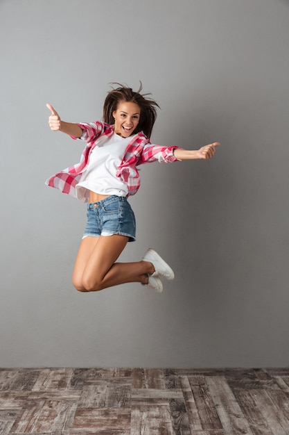 Full-length portrait of pretty jumping woman in casual wear, showing thumb up gesture