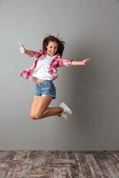 Full-length portrait of pretty jumping woman in casual wear, showing thumb up gesture