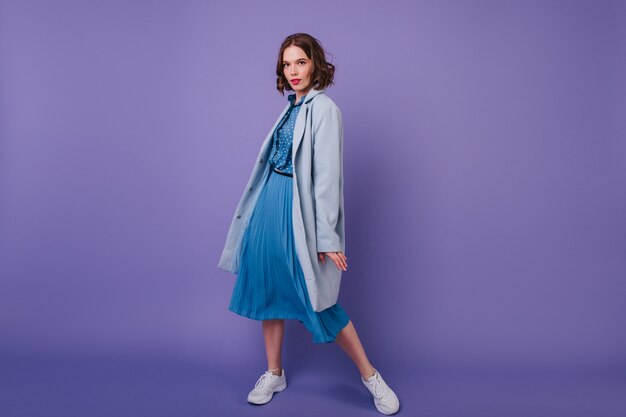 スニーカーとコートの洗練された女の子の全身像。紫色の壁に喜んでポーズをとる青い服を着た恍惚とした若い女性。