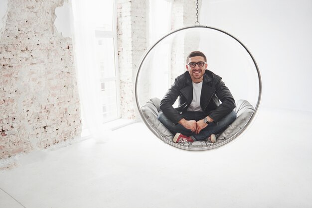 Full-length portrait of relaxed hipster man in casual sitting on hanging chair bullet and smiling on camera isolated over white