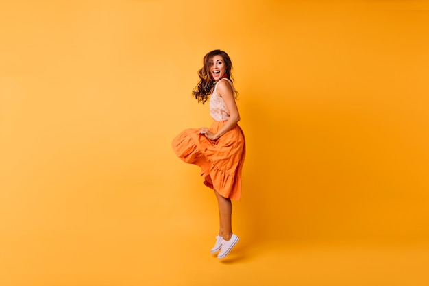 Full-length portrait of romantic beautiful lady in orange skirt. Stylish carefree girl jumping on yellow.