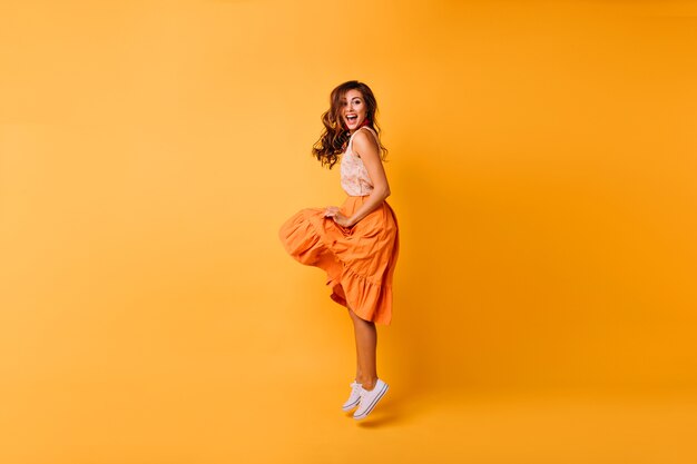オレンジ色のスカートのロマンチックな美しい女性の全身像。黄色にジャンプするスタイリッシュなのんきな女の子。