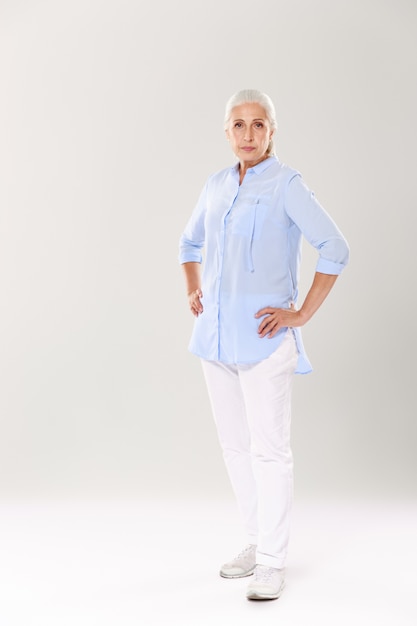 Full-length portrait of serious old lady in blue shirt and white pants, standing with hands on her waist,