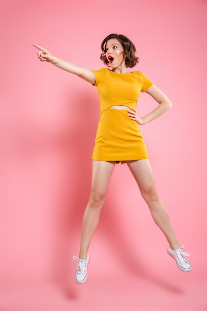 Full length portrait of shocked charming brunette woman with hand on her waist pointing with finger, looking aside while jumping over pink 