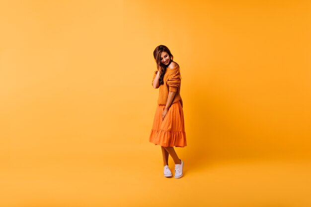 Полнометражный портрет застенчивой кавказской девушки, стоящей на желтом. Крытый снимок очаровательной девушки в оранжевом вязаном свитере и юбке.