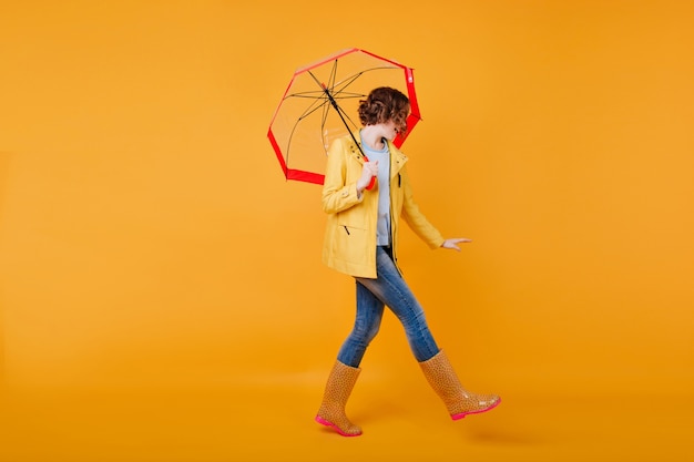 Full-length portrait of slim girl in funny rubber shoes dancing with umbrella. curly brunette lady having fun during photoshoot in autumn outfit