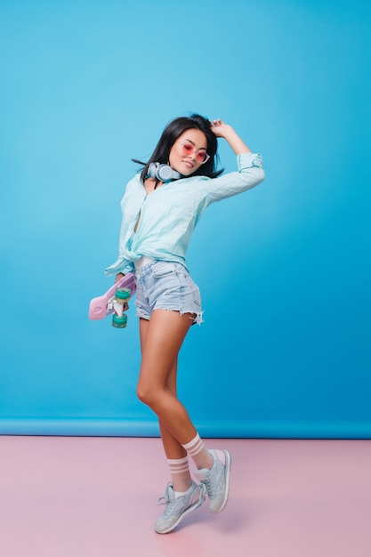 Full-length portrait of slim hispanic woman expressing happy emotions. Sensual latin girl with tanned skin in street outfit dancing with hand up.