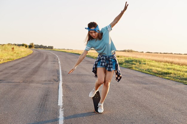 날씬한 스포티 여성이 스케이트보드 위에서 트릭을 하고, 혼자 활동적인 시간을 보내고, 거리에서 야외에서, 팔을 들고, 흥분한 표정으로 내려다보는 전체 길이 초상화.